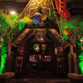 A vibrant Polynesian tiki bar in London featuring exotic cocktails, colorful lanterns, and bamboo furnishings. A bartender serves a flaming Scorpion Bowl in a traditional tiki mug, surrounded by lush greenery.