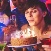Group of friends in a colourful bar as the birthday guest blows out candles on a cake while holding vibrant cocktails at Sugar Cane in Clapham.