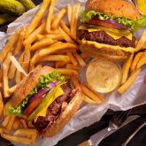 Overhead view of two gourmet burgers with melted cheese, lettuce, tomato, and fries on a wooden board, served with sauces at Sugar Cane Bar in London.