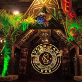 Exterior of The Sugar Cane Bar in London at night, featuring Tiki-style entrance, tropical palm trees, and colourful neon lighting.