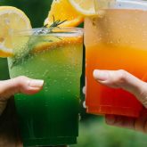 Two colourful summer cocktails garnished with lemon and orange slices, held up in a toast against a green outdoor background